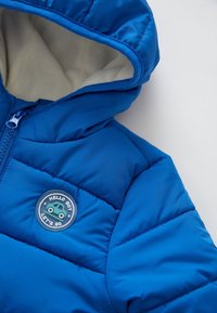 Niebieska ocieplana kurtka dla niemowląt z kapturem, beżowe polarowe wykończenie, zamek z przodu oraz okrągła naszywka z samochodem i napisem "WITAJ CHŁOPIE, CHODŹMY."