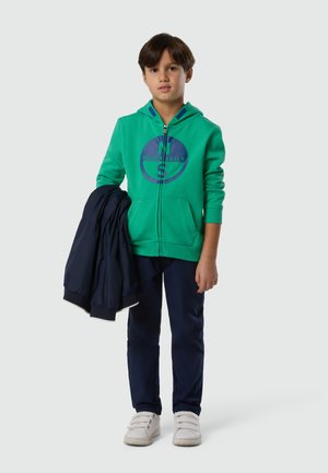 Junge mit braunen Haaren, der einen grünen Kapuzenpullover mit Reißverschluss und marineblaue Hose trägt, hält eine marineblaue Jacke in seiner linken Hand und steht vor einem einfarbigen Hintergrund.