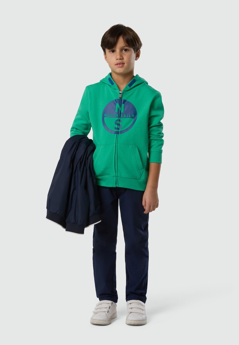 Garçon aux cheveux bruns portant un sweat à capuche vert zippé et un pantalon bleu marine, tenant une veste bleu marine dans sa main gauche, debout sur un fond uni.