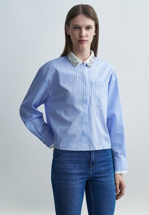 Žena oblečená v svetlomodrej blúzke s pruhmi a korálkovým bielym golierom, kombinovaná s tmavomodrými džínsami, stojaca pred jednoduchým sivým pozadím.