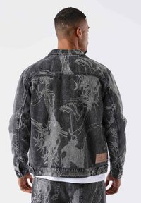 Chaqueta de mezclilla negra con un patrón floral texturizado en plata, cuello estándar y cierre de botones. Presenta un parche de cuero con marca en la parte posterior.