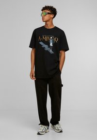 Svart t-skjorte med grafisk print og oransje tekst, kombinert med svarte cargo-bukser og mønstrede sneakers; modell iført grønne solbriller.
