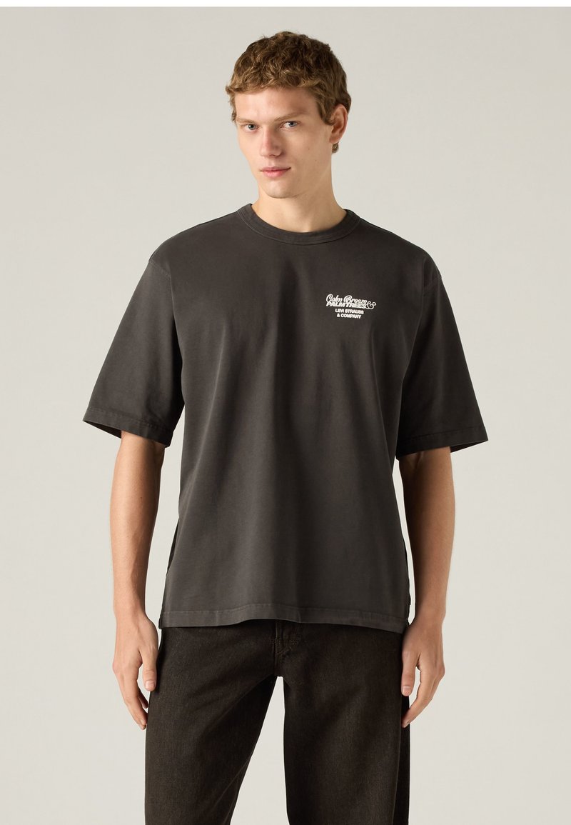 Young man wearing dark gray Levi Strauss t-shirt and black pants standing against a plain light background.