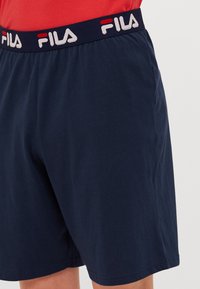 Námornícke modré šortky s elastickým pásom, na ktorých sú opakujúce sa biele a červené logo FILA, nosené s červeným vrchom.