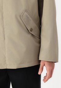 Casaco impermeável bege com um bolso com aba abotoada, textura suave e um corte ajustado. A marca está discreetamente estampada no bolso.