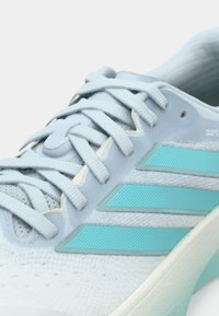 Zapatilla deportiva azul claro con parte superior de malla, tres franjas turquesa texturizadas, sistema de cordones planos y suela exterior de goma suave.