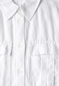 Camicia bianca in lino con colletto a punta, due tasche sul petto e chiusura a bottoni. Presenta una texture morbida e dettagli di cucitura discreti.