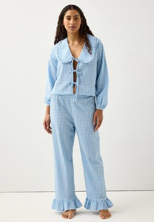 Kvinde iført blåt og hvidt gingham-nattøjssæt med stor krave, knaplukninger foran, lange ærmer og flæsekant på buksebenene, stående barfodet.