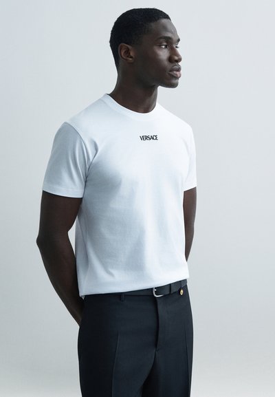 Weißes Baumwoll-T-Shirt mit kurzen Ärmeln und einem schwarzen "VERSACE"-Logo auf der Brust. Kombiniert mit einer dunklen Hose und einem Gürtel.