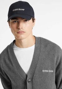 Cardigan grigio a maglia con scollo a V, dotato di una chiusura con un bottone e del testo ricamato "GUESS JEANS". Abbinato a un cappellino scuro.