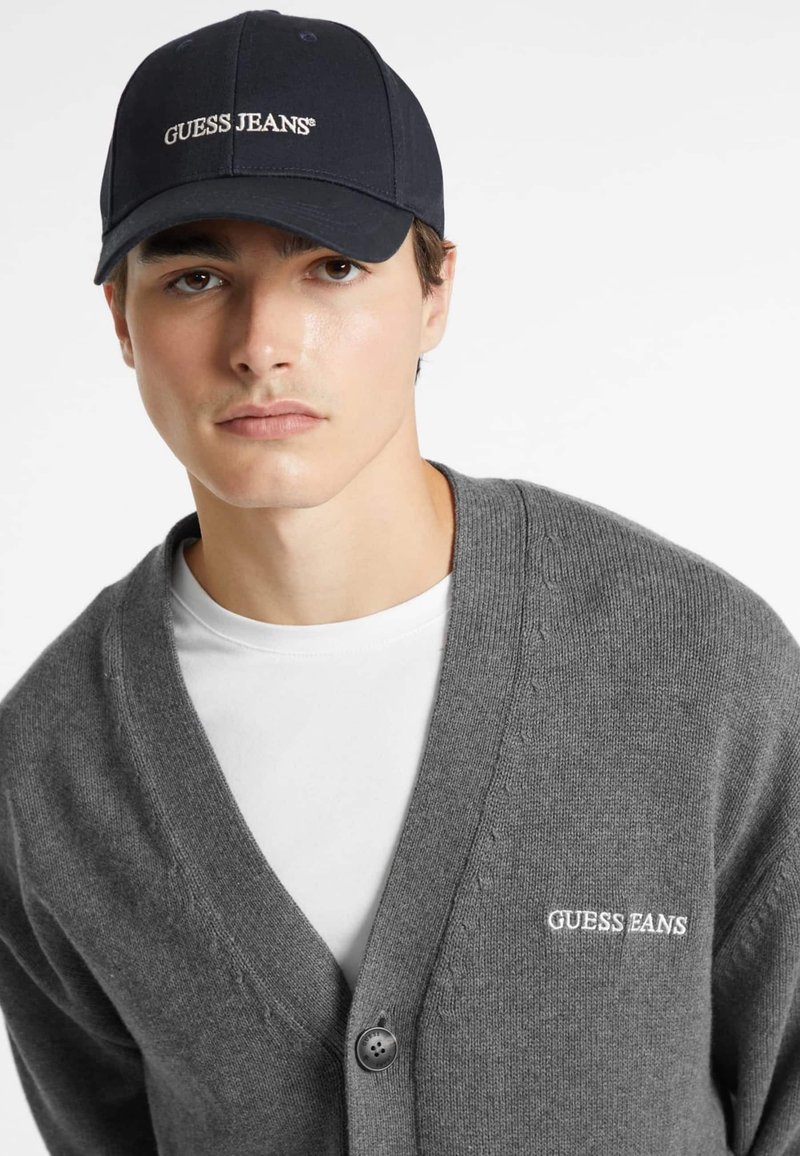 Cardigan grigio a maglia con scollo a V, dotato di una chiusura con un bottone e del testo ricamato "GUESS JEANS". Abbinato a un cappellino scuro.