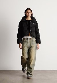 Veste courte en duvet noire, texture matelassée, fermeture par boutons, associée à un jean oversize délavé et des chaussures grises, le tout sur un fond neutre.