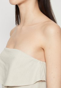 Ανοιχτό καφέ strapless τοπ με φαρδιά ρέουσα αφέλεια, φτιαγμένο από υλικό με υφή, αποκαλύπτοντας τη λεία επιδερμίδα κατά μήκος του λαιμού και του ώμου.