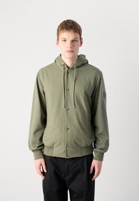 Joven vestido con una sudadera verde oliva con botones y cordones, y pantalones negros, de pie frente a un fondo claro y simple.