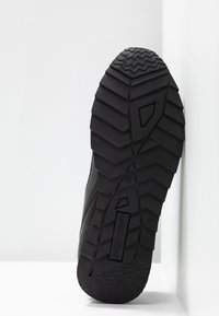Black rubber shoe sole with deep treads and angular patterns. Features a branded section and a distinct edge along the outer rim.