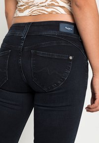 Närbild av en person som bär mörkblå Pepe Jeans med bakfickor, ihop med en topp i brunt och vitt mönster.