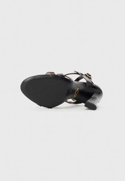 Black heeled sandal with a smooth leather upper, crossed strap design, gold accents, and a textured heel. Flat black sole for grip.