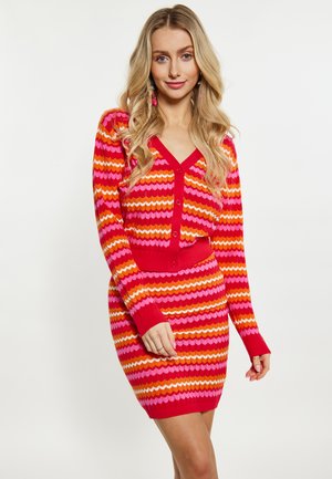 Femme aux longs cheveux blonds portant une robe en tricot rayée rouge, orange et rose avec des boutons, debout devant un fond clair uni.