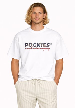 Mann mit schulterlangem blondem Haar trägt ein weißes T-Shirt mit der Aufschrift "POCKIES a couch couture company" und hellbeige gestreifte Hose mit Kordelzug.
