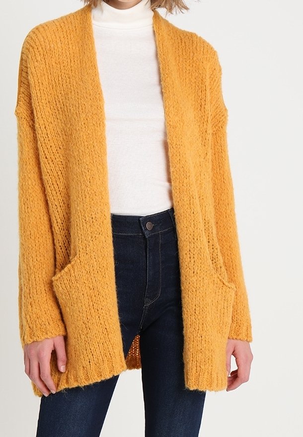 Mustard yellow knitted cardigan with open front, long sleeves, and front pockets. Worn over a white turtleneck and dark jeans.