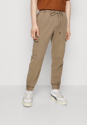 Beige cargobroek met een elastische tailleband en trekkoord, voorzien van twee zijzakken en elastische manchetten. Gekleed met witte sneakers.