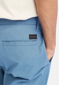 Niebieskie spodnie z materiału z tylną kieszenią i czarną metką z logo Timberland. Materiał ma gładką fakturę i dopasowany krój.