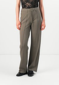 Pantalons évasés en tissu vert olive, dotés d'une taille à cordon, de plis et d'une texture lisse. Portés avec des chaussures noires.
