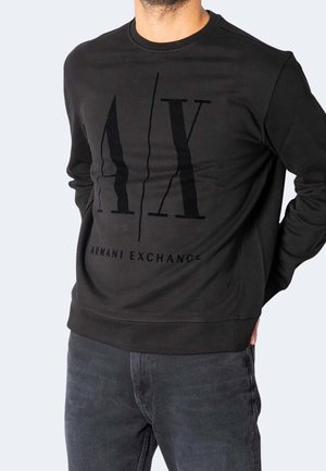 Mężczyzna w czarnej, długorękawowej bluzie Armani Exchange z dużym logo "A|X" i czarnych dżinsach, ręce za plecami.