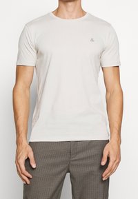 Jasno beżowa koszulka z krótkim rękawem, okrągłym dekoltem i małym logo na lewej piersi, wykonana z miękkiego, gładkiego materiału.