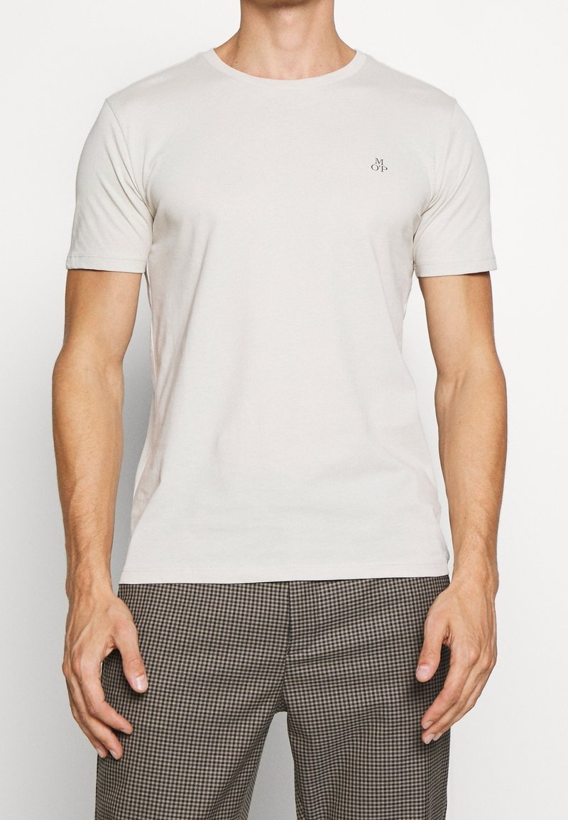 Jasno beżowa koszulka z krótkim rękawem, okrągłym dekoltem i małym logo na lewej piersi, wykonana z miękkiego, gładkiego materiału.