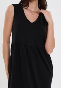 Czarna sukienka bez rękawów z dekoltem w serek, z marszczonym stanem i gładką fakturą. Częściowo widoczne warstwowe naszyjniki.