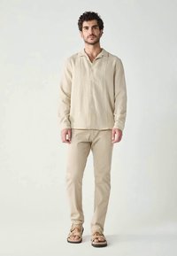 Chemise beige clair à boutons avec texture verticale, associée à un pantalon beige assorti et des sandales marron. Col simple et coupe décontractée.