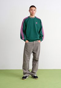 Green fleece sweatshirt with pink stripes, logo on the chest, paired with gray corduroy pants and navy sneakers. Plain background.