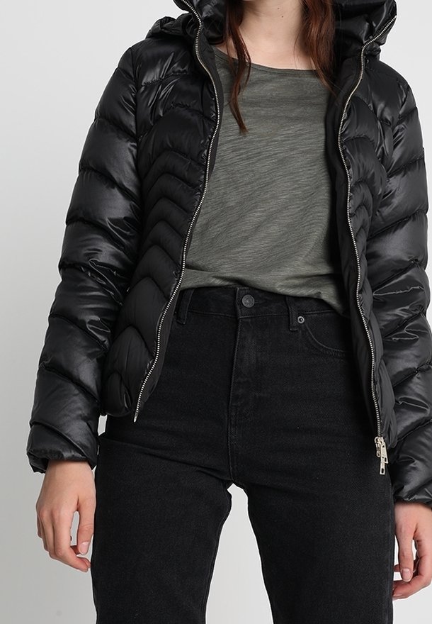 Femme portant une veste noire matelassée brillante avec une capuche, un t-shirt gris et un jean noir taille haute, debout les bras détendus.