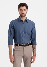 Camicia button-up in tessuto blu scuro, maniche lunghe arrotolate fino ai gomiti, taschino sul petto, abbinata a pantaloni beige chiaro e una cintura marrone.