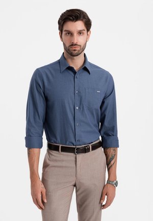 Camisa de botones de tela azul oscura, con mangas largas arremangadas hasta los codos, bolsillo en el pecho, combinada con pantalones beige claros y un cinturón marrón.