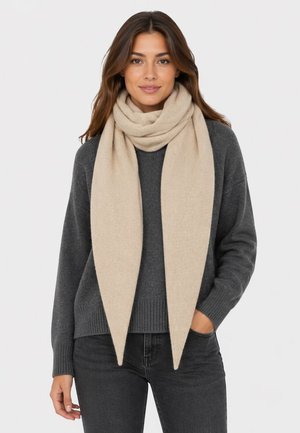 Woman wearing a beige scarf wrapped around her neck, dark gray sweater, and black jeans, with one hand in pocket, facing forward.