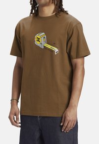 Brown cotton t-shirt featuring a graphic of a tape measure in gray, yellow, and black with the text "SIZE MATTERS". Short sleeves, relaxed fit.