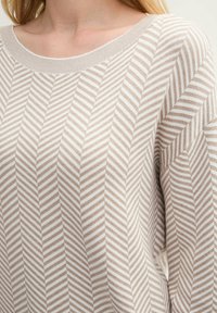 Beige gestreepte gebreide trui met een getextureerd zigzagpatroon, brede halslijn en een ontspannen pasvorm, die de zachte stof en diagonale lijnen accentueert.