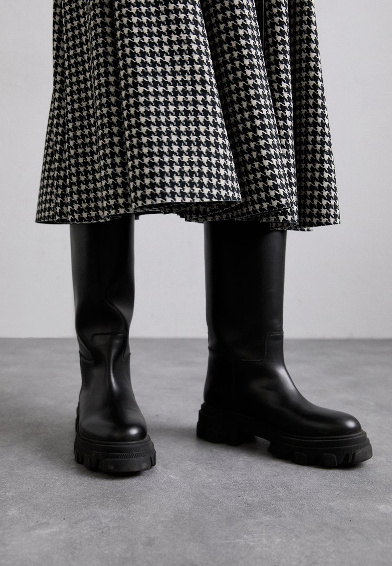 Črne usnjene gležnjarje do kolen z masivnimi podplati, ki se odlikujejo po gladki teksturi in minimalnem šivu, v kombinaciji z krilom v houndstooth vzorcu.