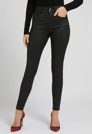 Femme portant un jean skinny noir taille haute et des talons hauts bordeaux, une main dans la poche, sur un fond gris uni.