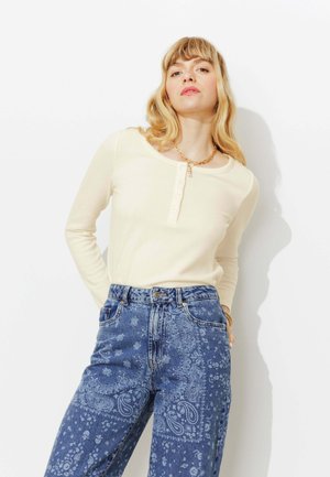 Femme aux cheveux blonds ondulés portant un haut henley crème à manches longues et un jean taille haute bleu à motif cachemire, debout devant un fond blanc.