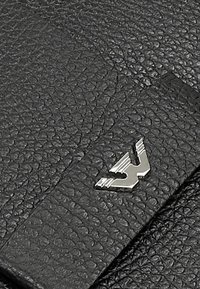 Czarny materiał skórzany o fakturowanej powierzchni, z akcentem w postaci srebrnego metalowego logo. Projekt zawiera detal z przeszywanymi brzegami.