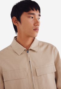 Beige canvas jack met een kraag, rits aan de voorkant en twee borstzakken. Heeft een horizontale textuur en een losse pasvorm.