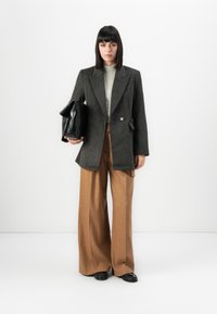 Femme se tenant devant un fond blanc, portant un blazer gris, un haut rayé, un pantalon large marron, des chaussures noires, tenant un grand sac à main noir.