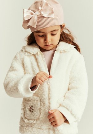 Junges Mädchen trägt eine rosa Strickmütze mit Schleife und eine weiße, flauschige Jacke, konzentriert darauf, die Jacke zuzumachen.