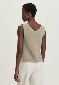 Top beige a maglia senza maniche con scollo a V sul retro e motivo testurizzato, indossato con pantaloni larghi di colore chiaro.
