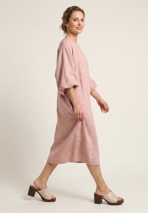 Frau trägt ein locker sitzendes, pinkes Midikleid und beige Sandalen mit Absatz vor einem schlichten hellen Hintergrund.
