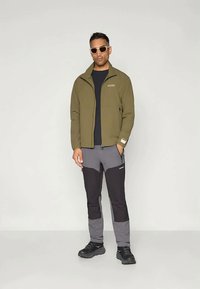 Olive green lightweight jacket with zippered pockets, paired with gray and black pants featuring color blocking. Sturdy black shoes complete the look.