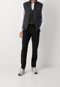 Gevoerde zwarte vest over een witte lange mouwen shirt, grijze gebreide trui, zwarte broek en grijze sneakers, met een rits aan de voorkant en logo-detail.